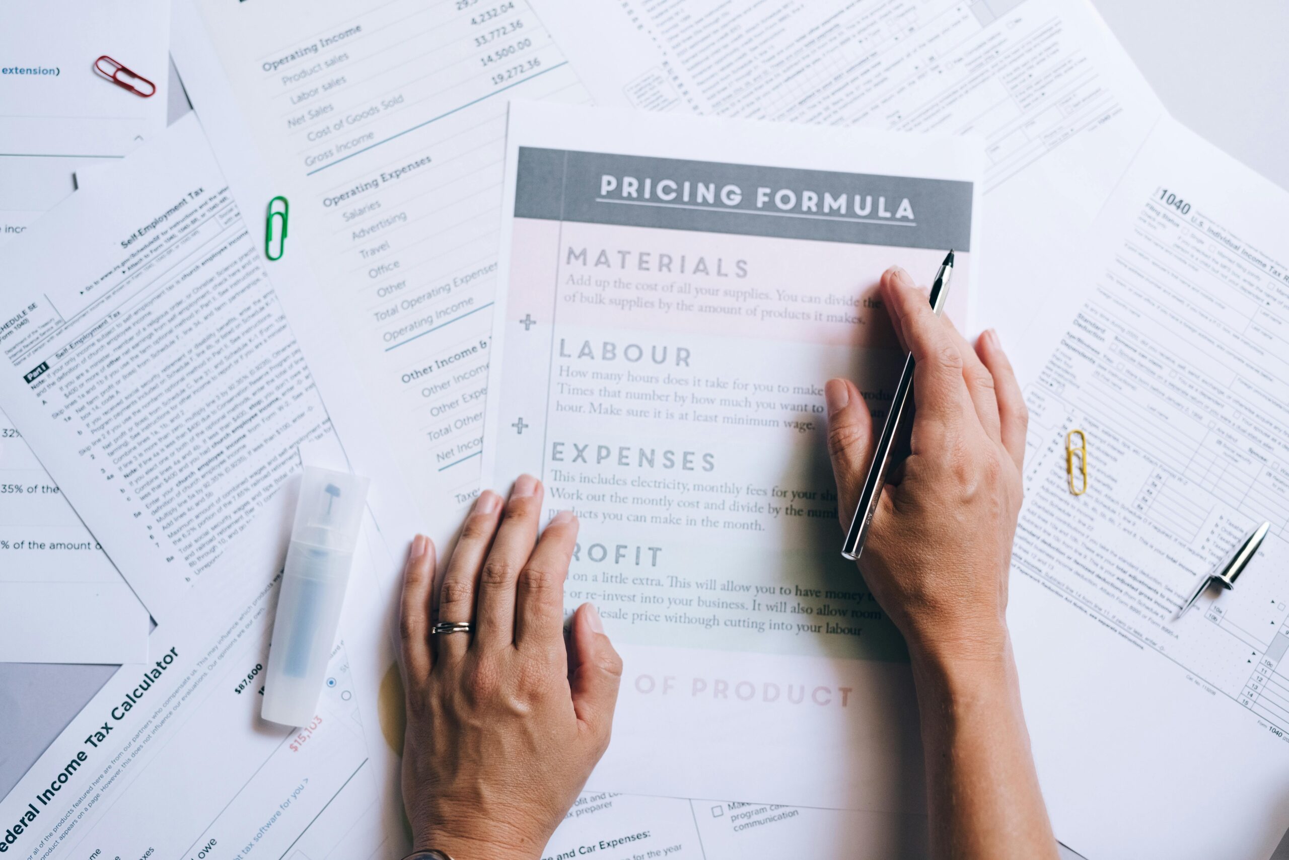 Hands organizing business documents and pricing formula papers on an office desk.